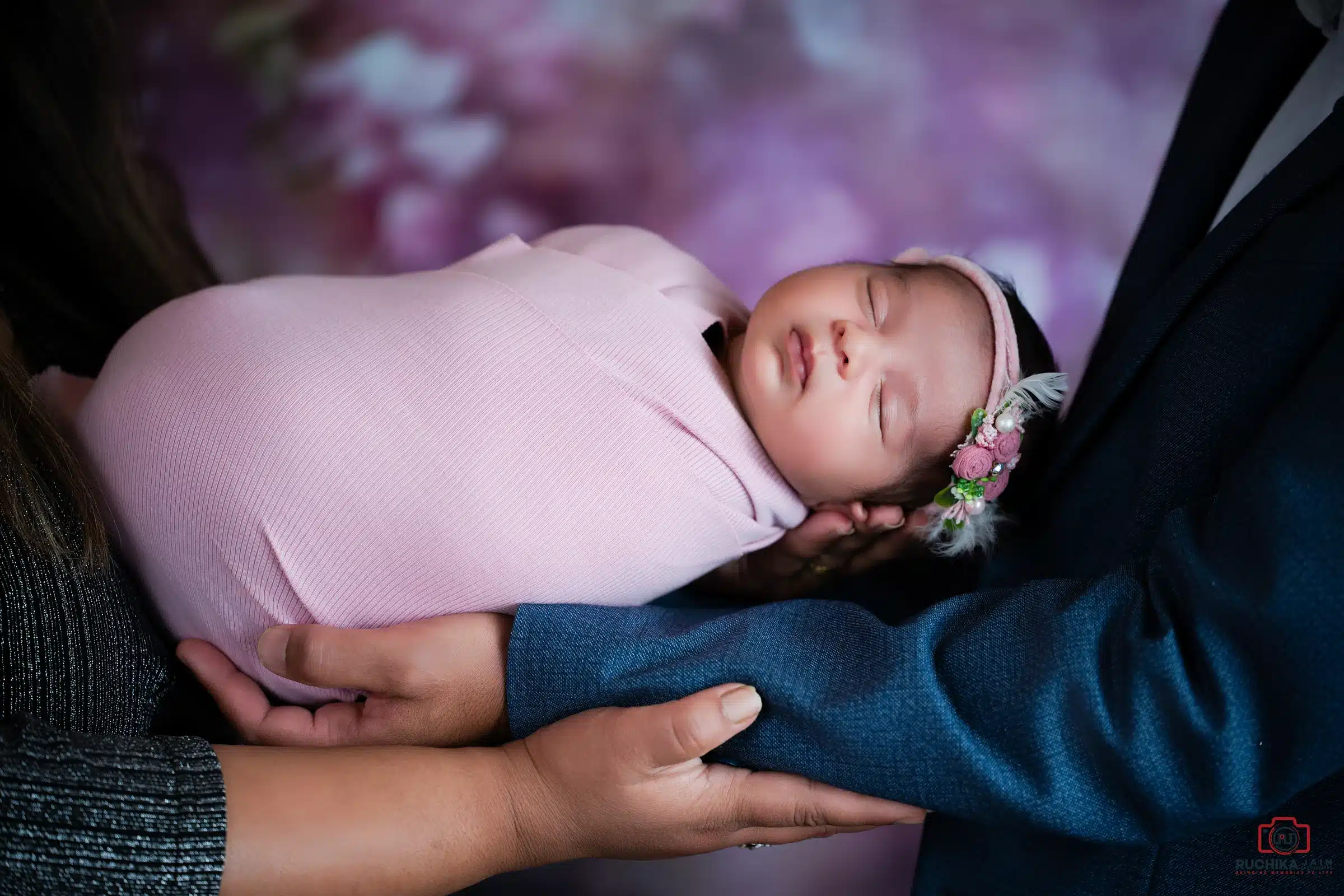 Newborn photography close-up of a sleeping baby wrapped in pink and held in parents’ arms.